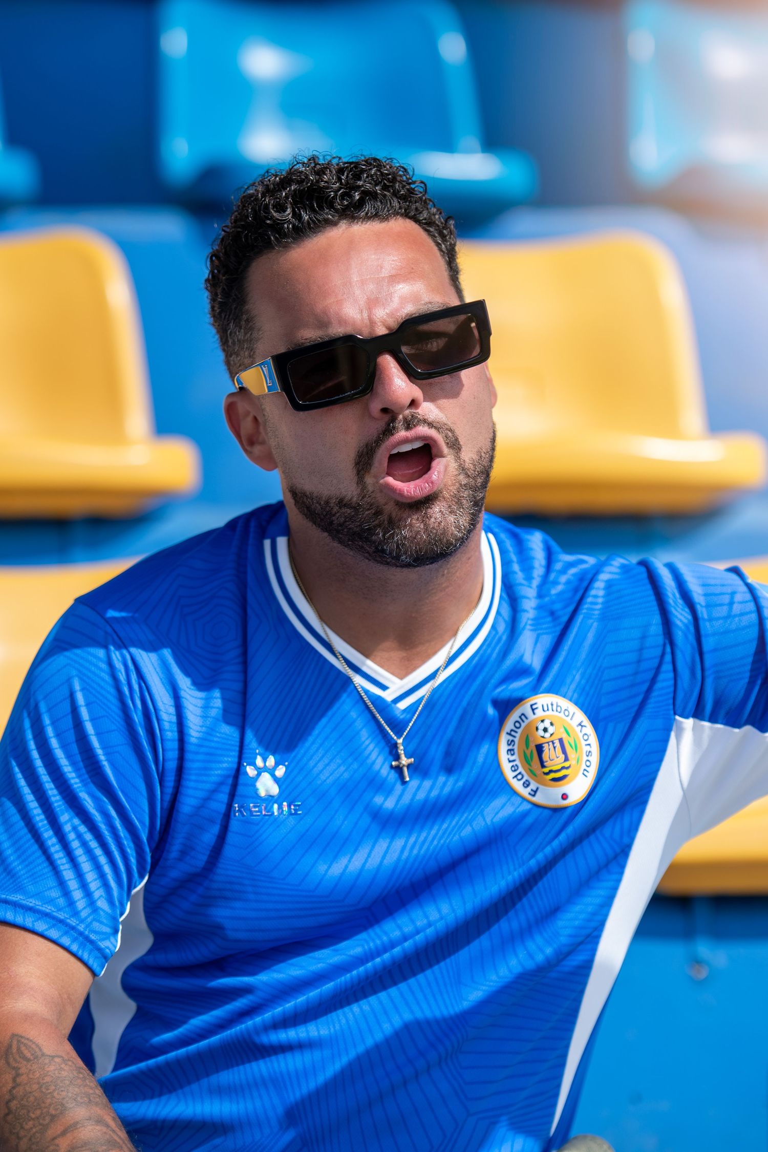 Man in blue soccer jersey and sunglasses sitting in stadium seats on a sunny day.