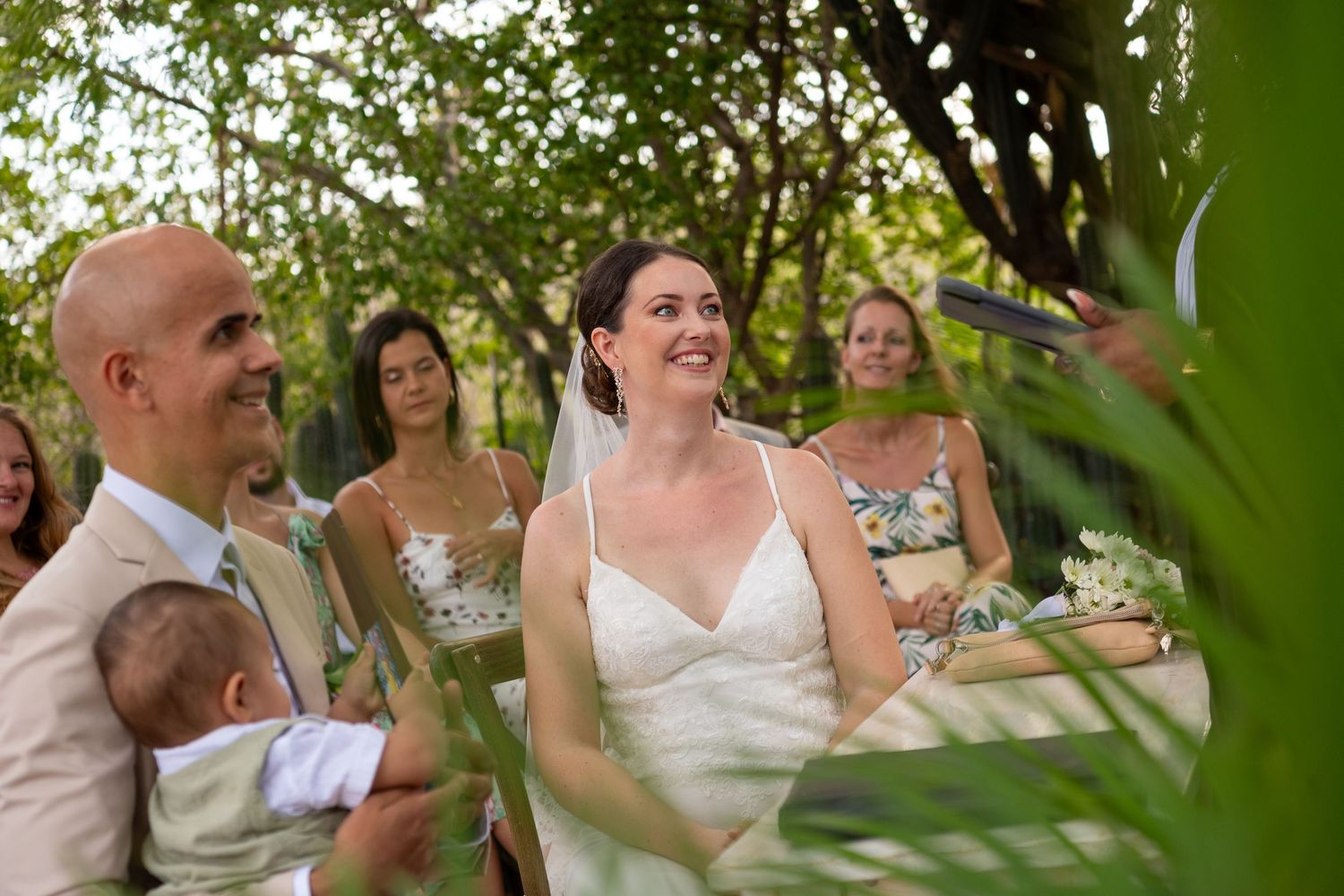 A wedding couple laughs joyfully while seated outdoors under green trees and white flowers.