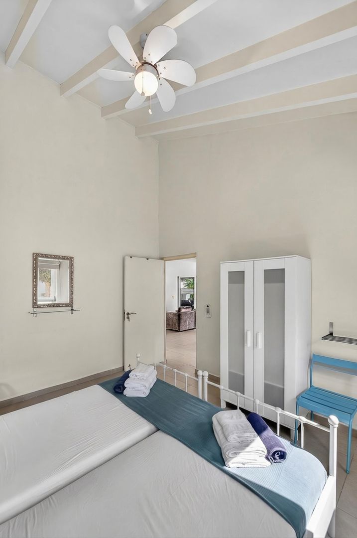 Bright white bedroom with high beamed ceiling, ceiling fan, metal bed frame, teal bedding, and white wardrobe.