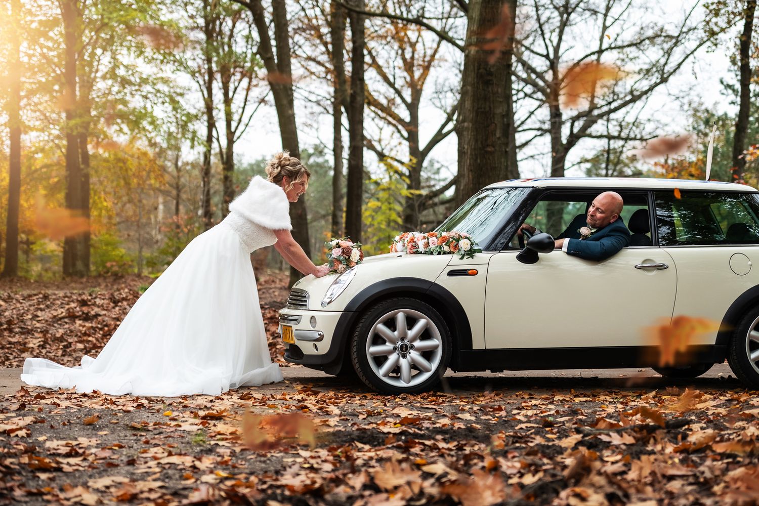A bride in a white wedding dress playfully checks under the hood of a white Mini Cooper on an autumn day covered in leaves.