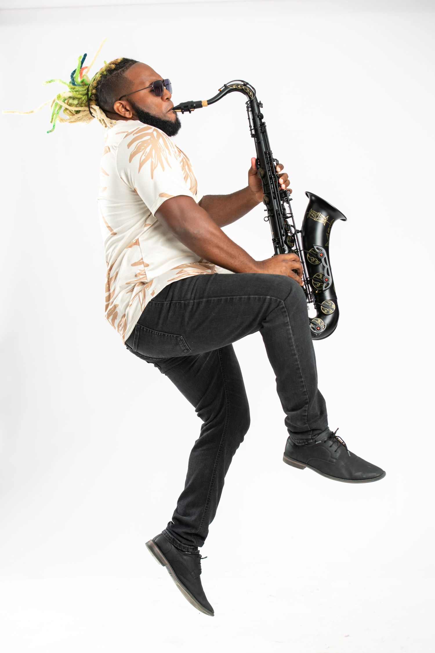 Man with dreadlocks jumping while playing black saxophone, wearing sunglasses and casual outfit on white background.