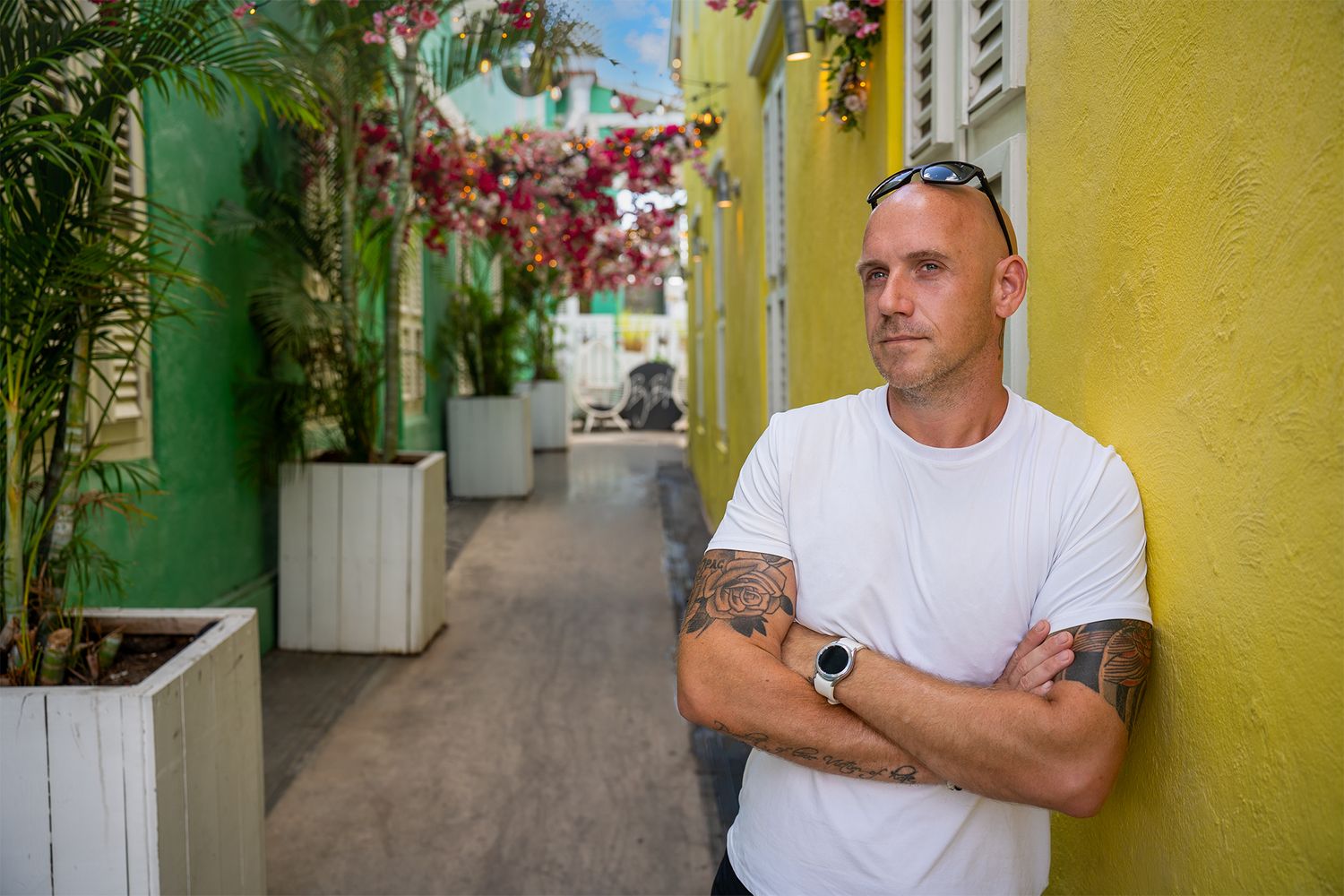 Person in white t-shirt stands against vibrant yellow wall with tropical plants and colorful architecture.