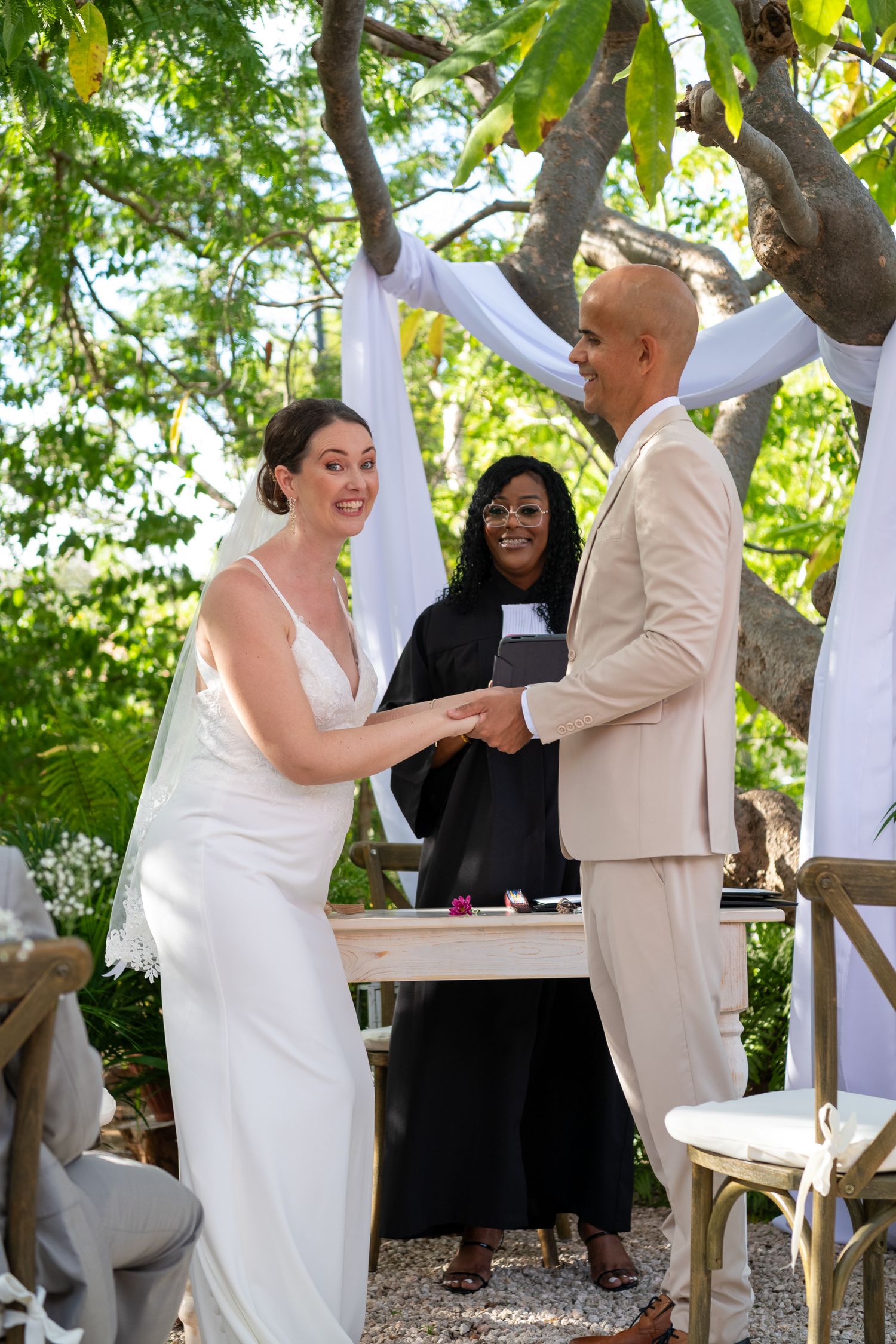 A wedding ceremony takes place outdoors under green trees as a couple exchanges rings with an officiant present.
