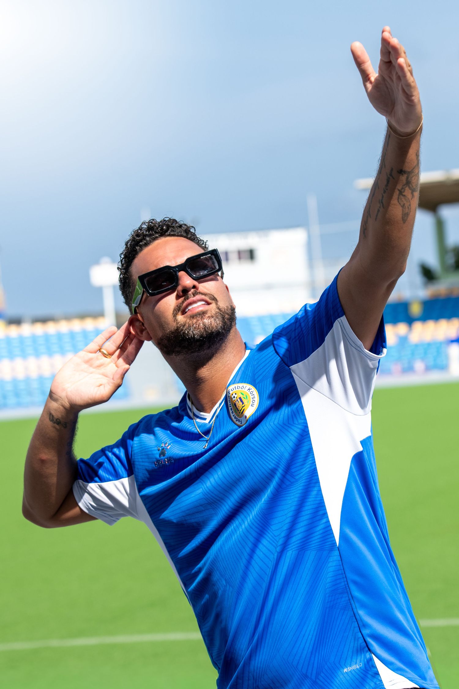 A man in a blue soccer jersey and sunglasses poses with arms raised at a stadium on a sunny day.