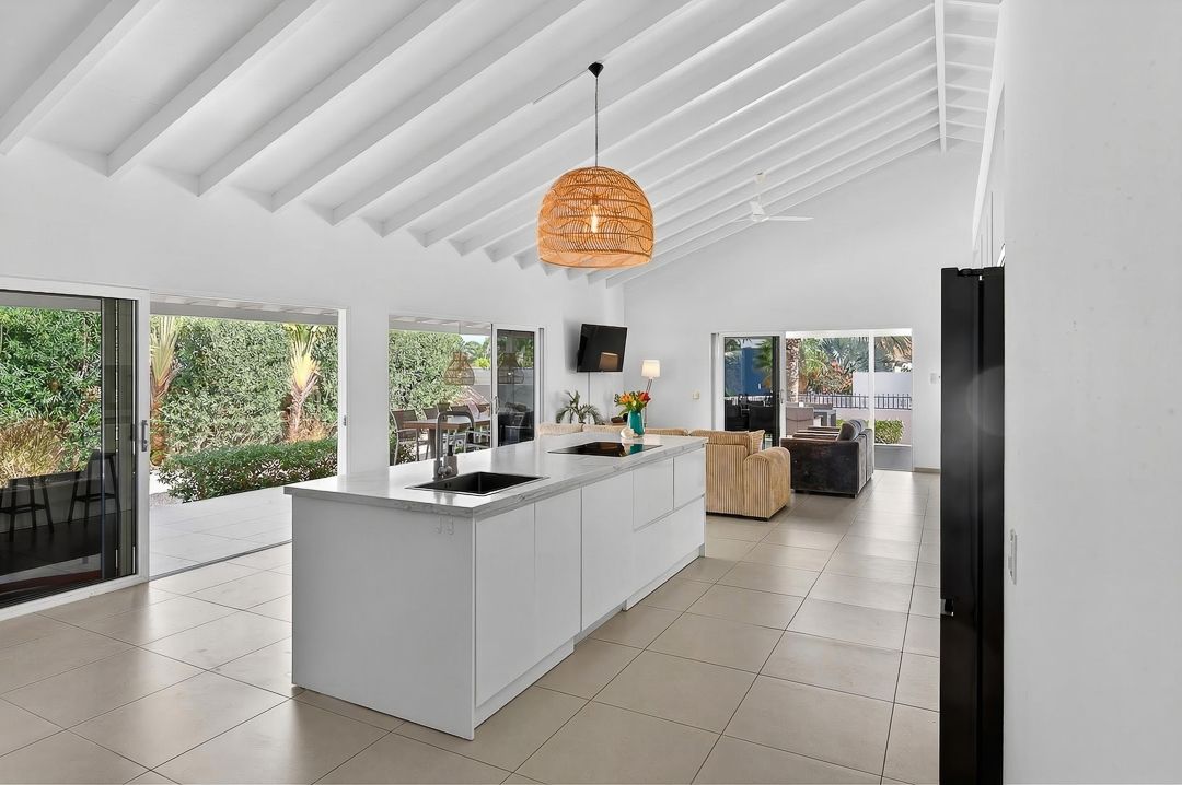 Modern open-plan kitchen with white cabinetry, island bench, rattan pendant light, and floor-to-ceiling windows overlooking lush greenery.