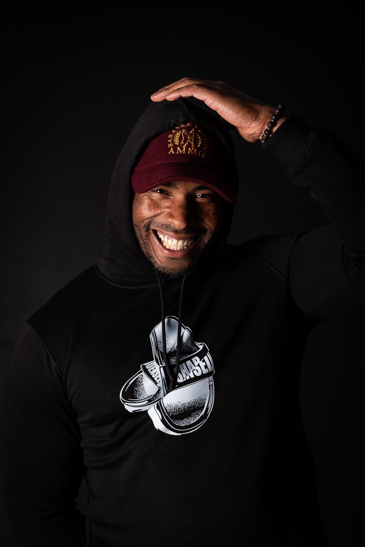 Portrait of person in black hoodie with sneaker graphic and burgundy cap posing against dark background with bright smile.