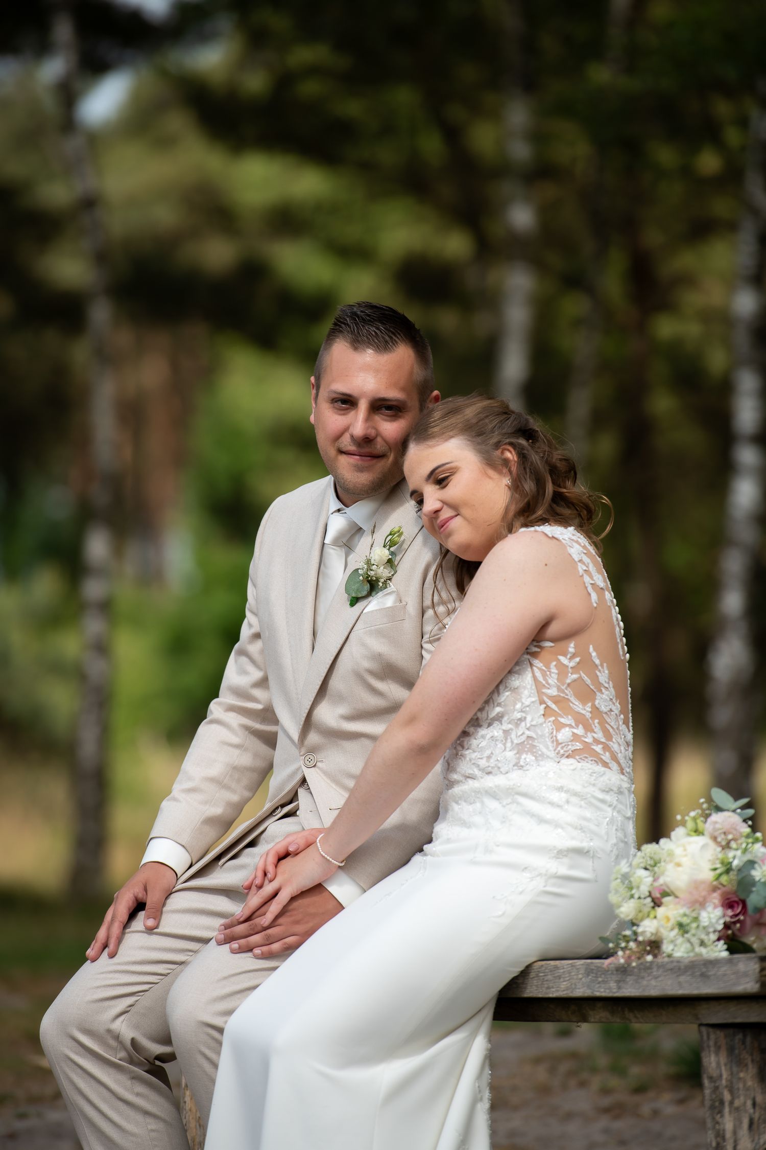 A romantic wedding couple sits together on a rustic bench in a wooded outdoor setting.