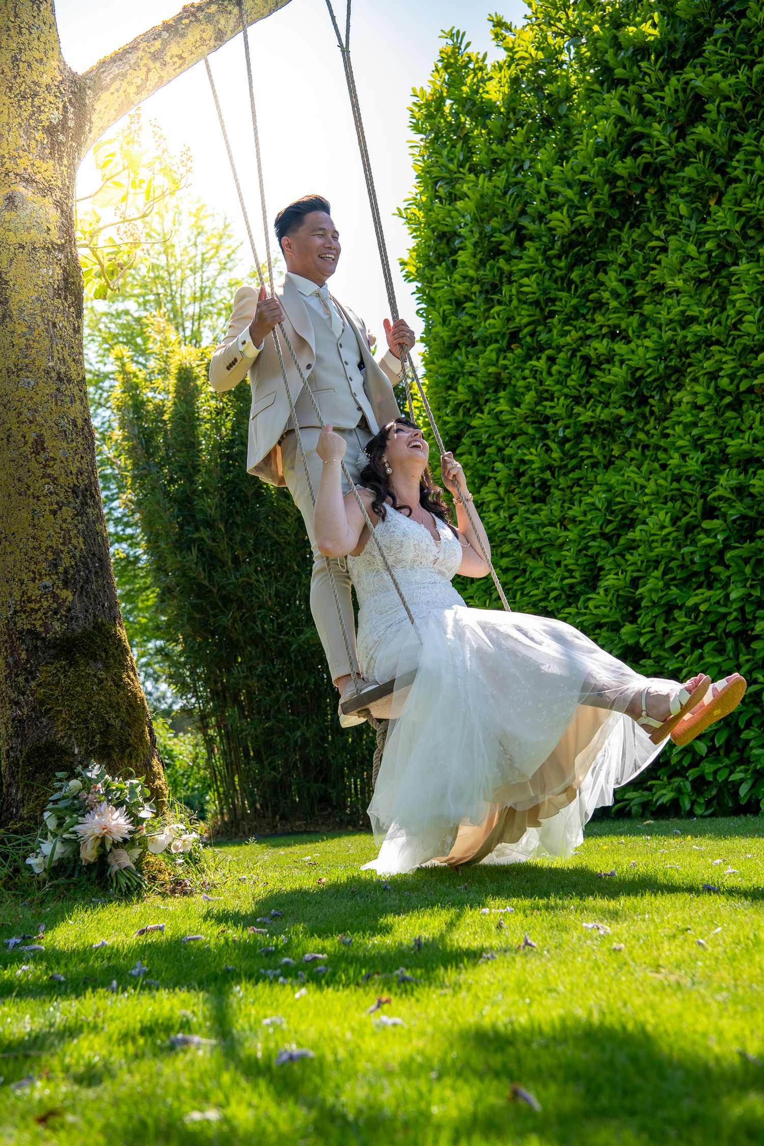 A couple in wedding attire swings together on a garden swing against a backdrop of lush green hedges and sunlight.
