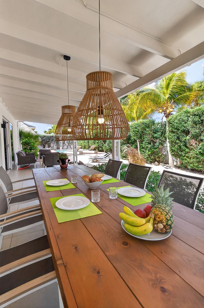 Outdoor dining area with wooden table, wicker pendant lights, fresh fruit, and tropical garden views under a covered patio.