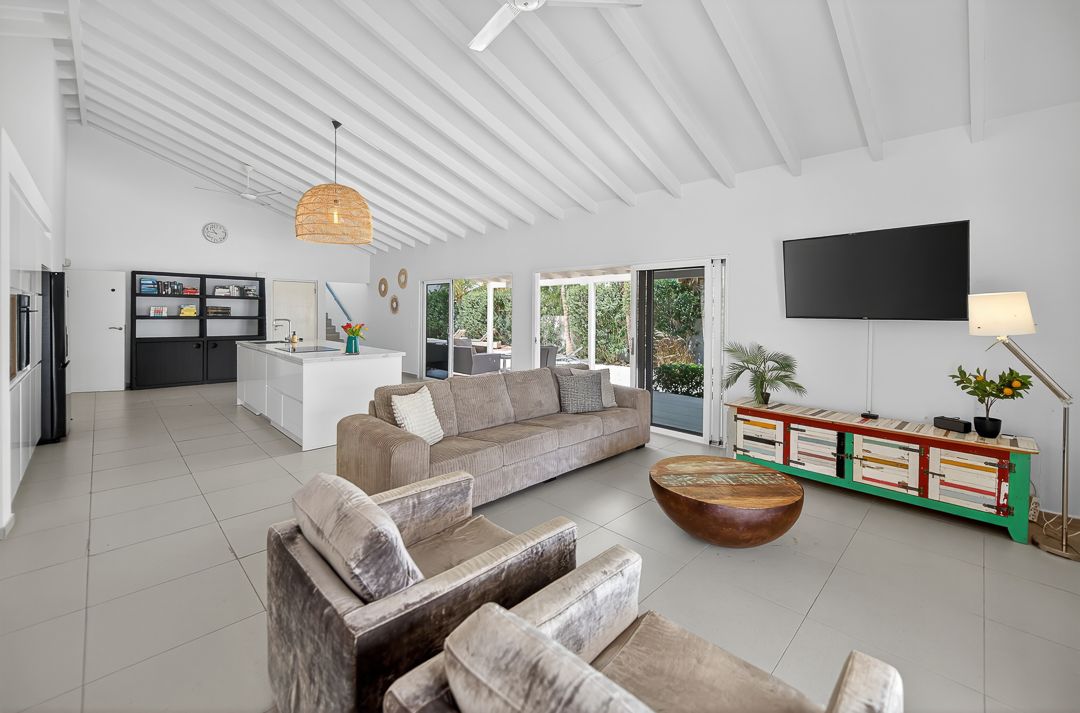 Bright open-plan living room with vaulted white ceilings, neutral sofas, wall-mounted TV, and sliding doors to lush greenery.