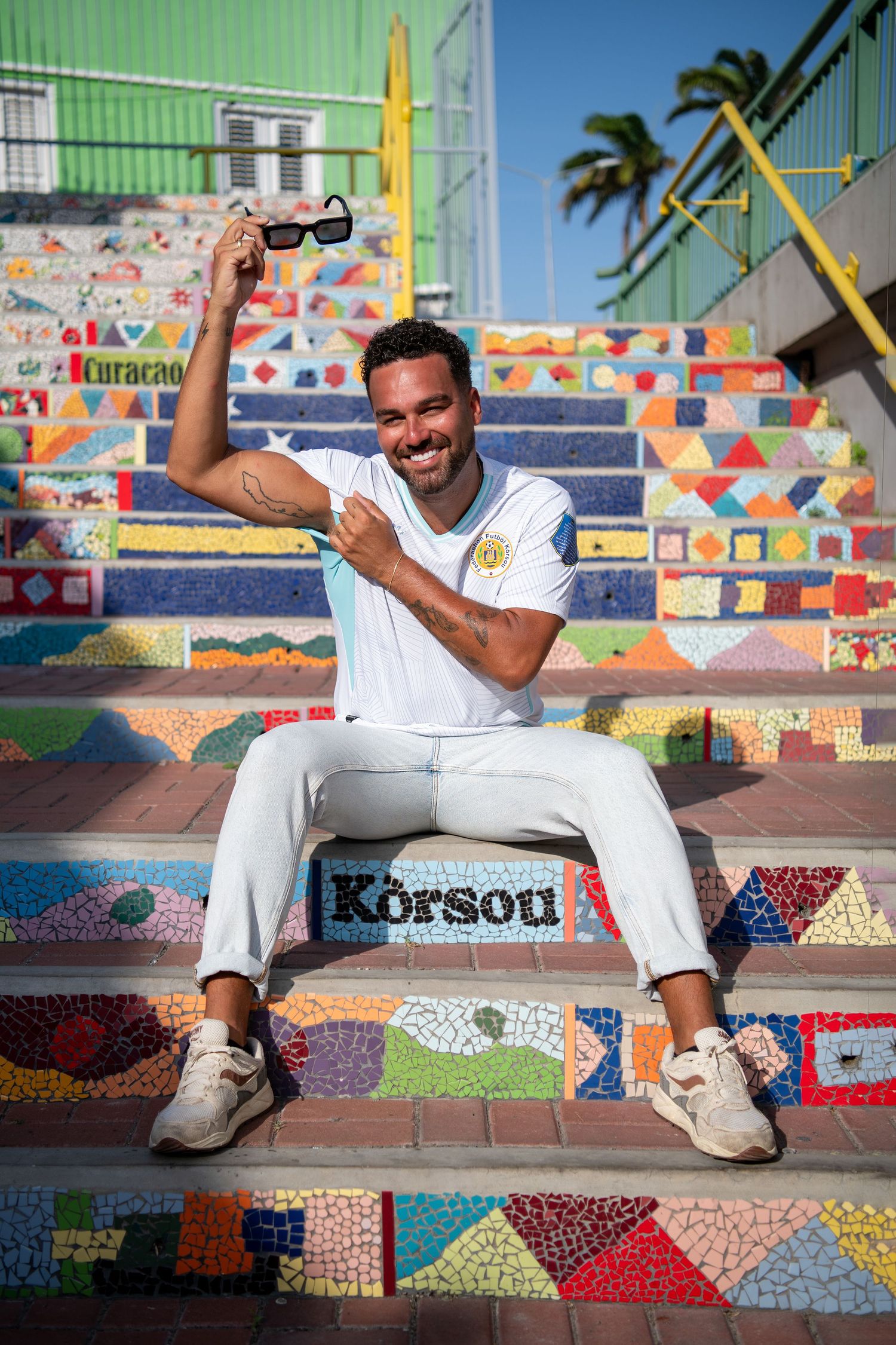 Smiling man in white outfit sitting on colorful mosaic steps in Curacao, holding sunglasses.