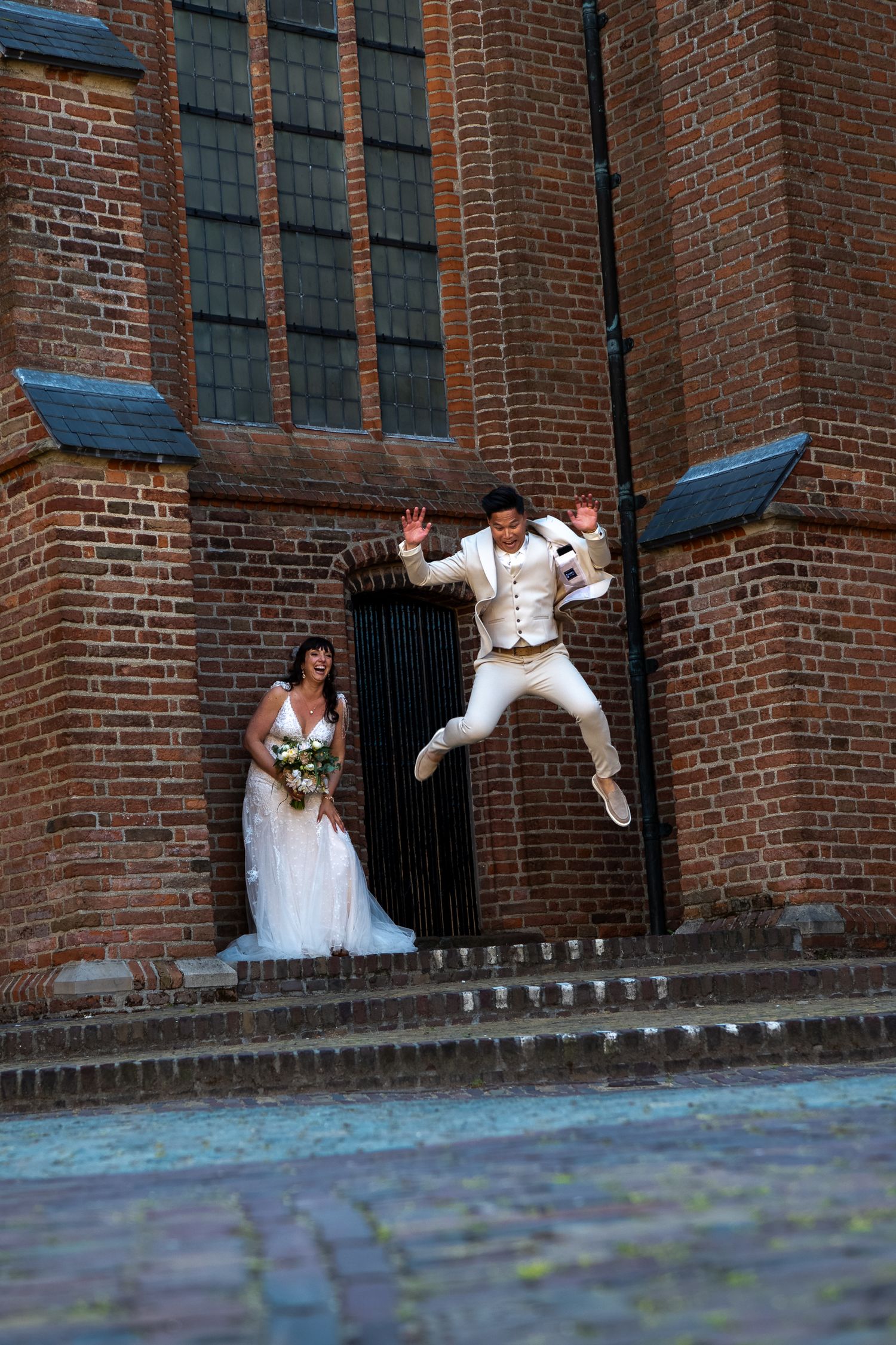 A couple poses for a wedding photo outside a brick church with one person jumping excitedly on the steps.