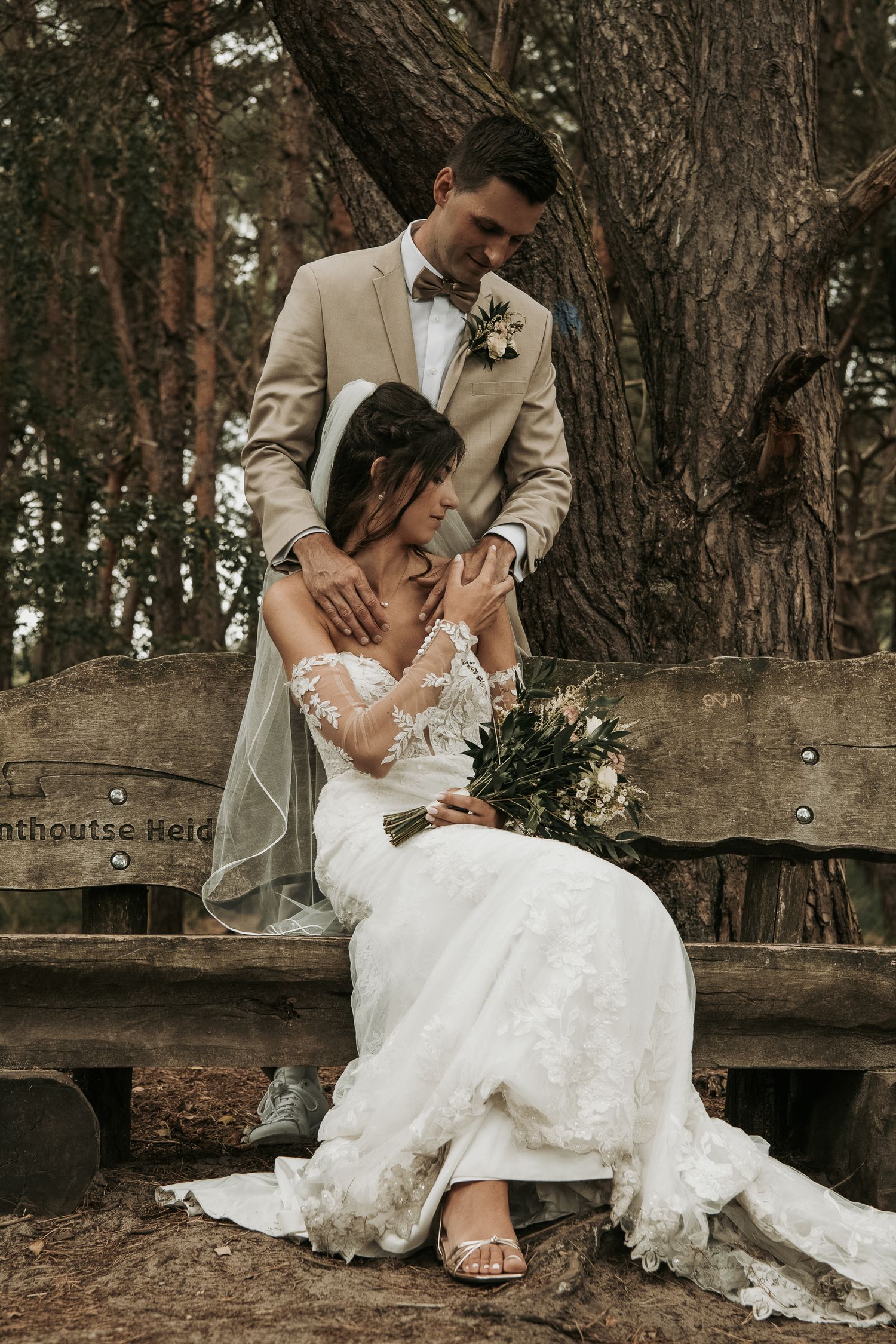 A romantic wedding couple poses together on a rustic wooden bench surrounded by tall trees in a forest setting.