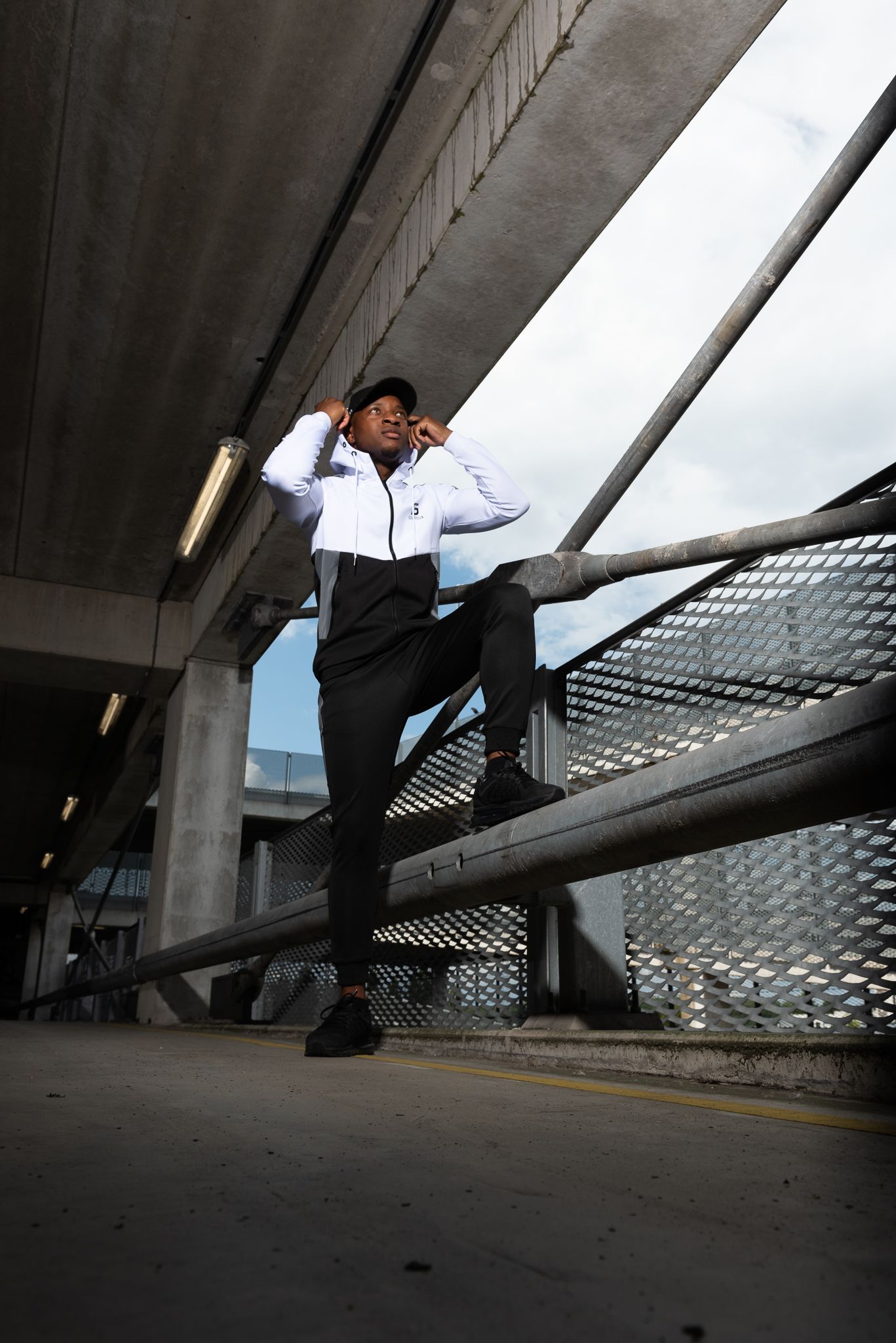 A person in black and white streetwear poses dramatically on an urban railing beneath a concrete overpass structure.