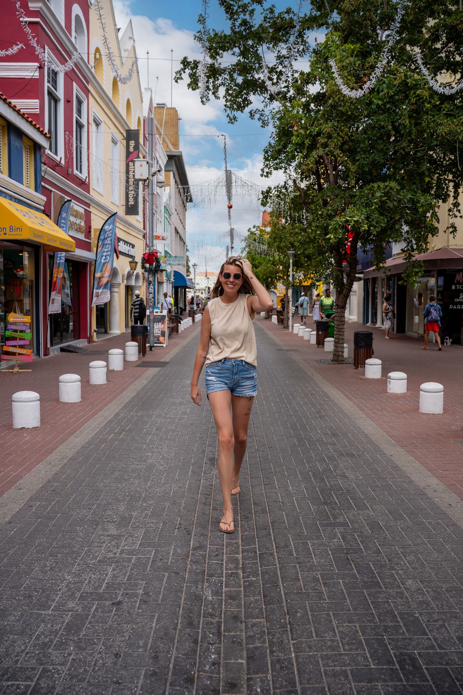 Woman in denim shorts and white tank top walking barefoot down a colorful pedestrian shopping street on a sunny day.