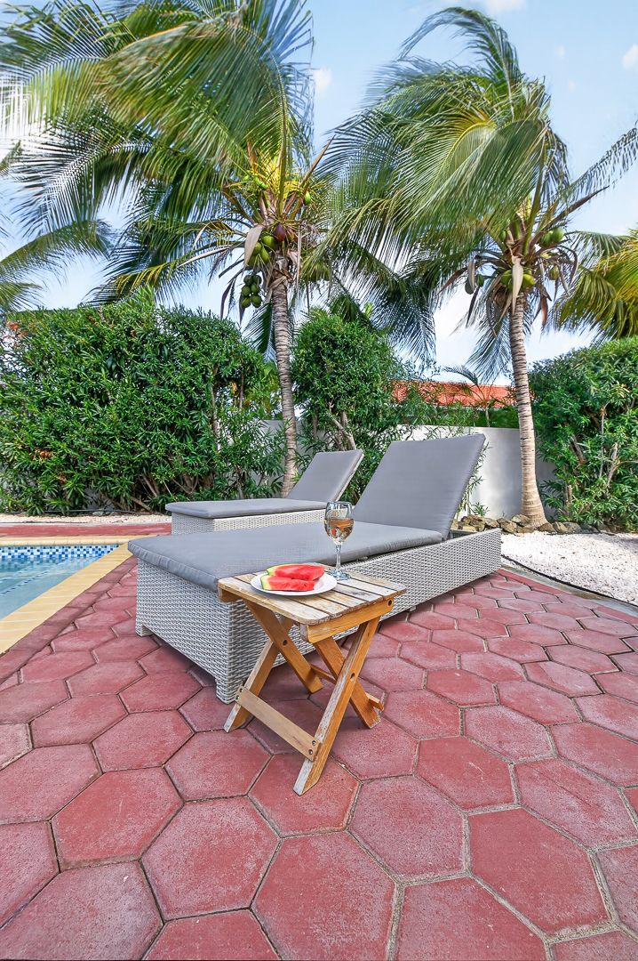 Two lounge chairs beside a small wooden table with wine and watermelon, set on red hexagonal pavers near a pool with palm trees.