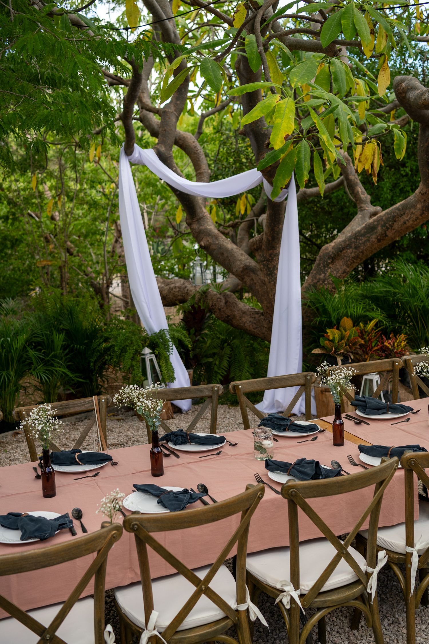Outdoor dining setup with white draping and place settings along a wooden railing under a leafy tree canopy.