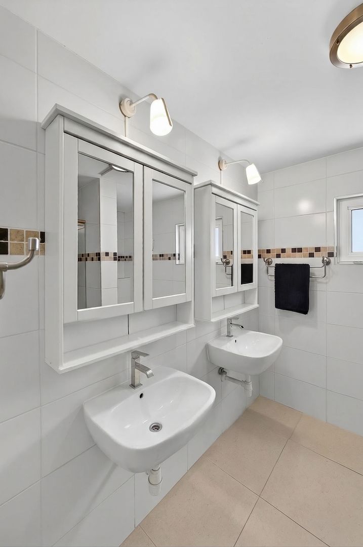 Modern white bathroom with wall-mounted sink, mirrored medicine cabinets, mosaic tile accents, and beige floor tiles.