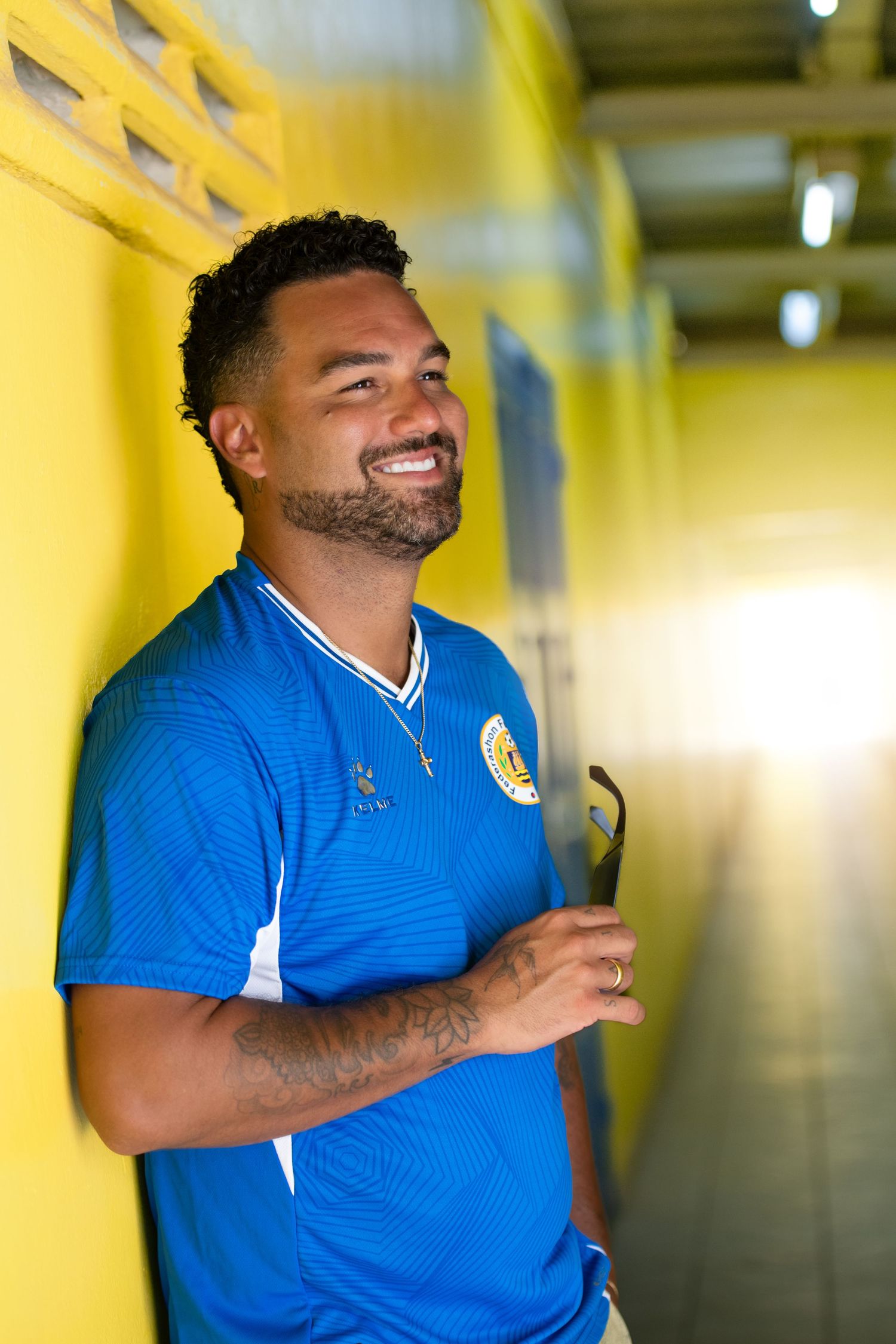Smiling man in blue soccer jersey leaning against yellow wall in a bright corridor, holding sunglasses.