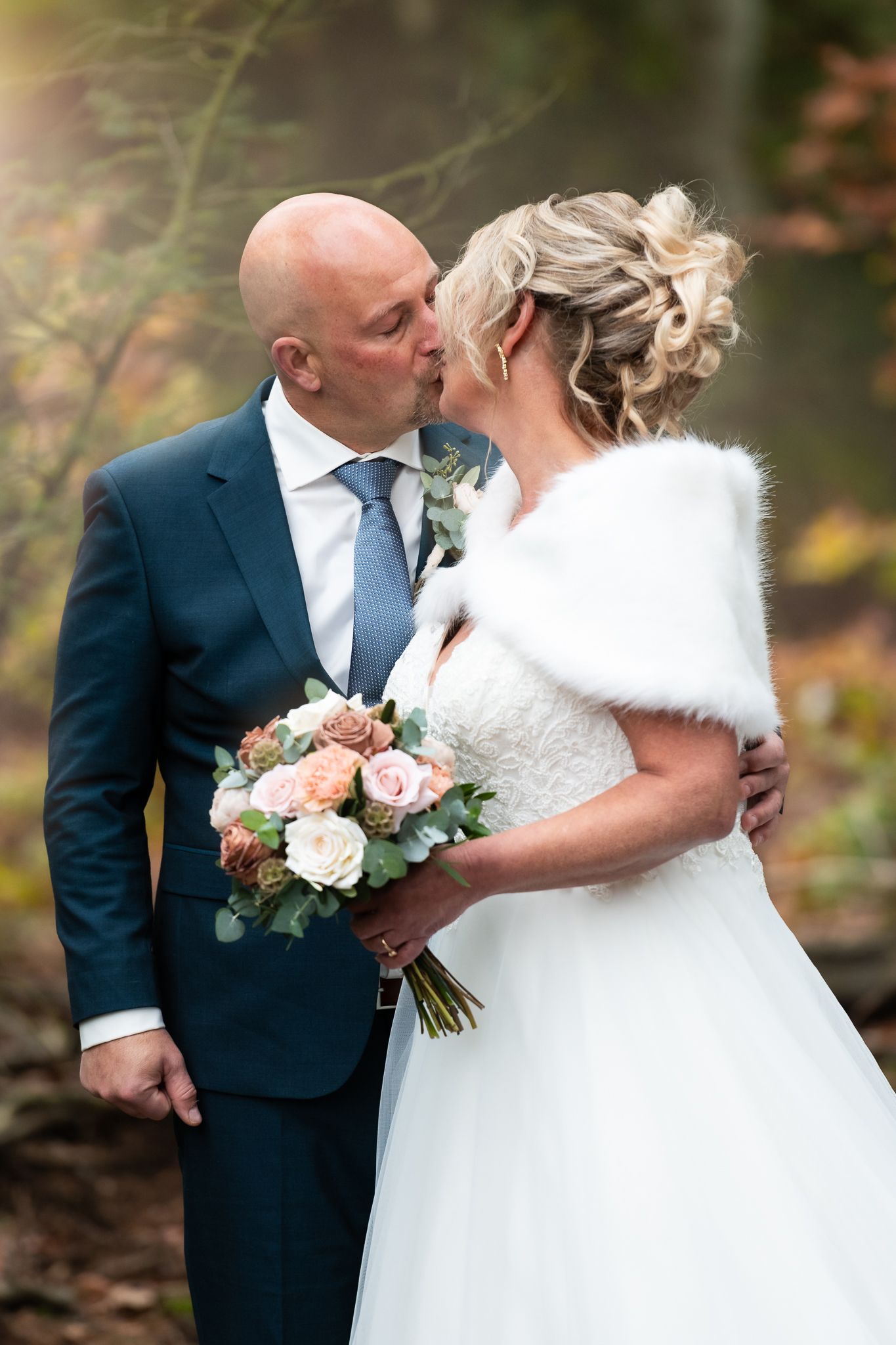 A romantic wedding couple embraces in an autumn garden setting with soft pink and white floral bouquet details.