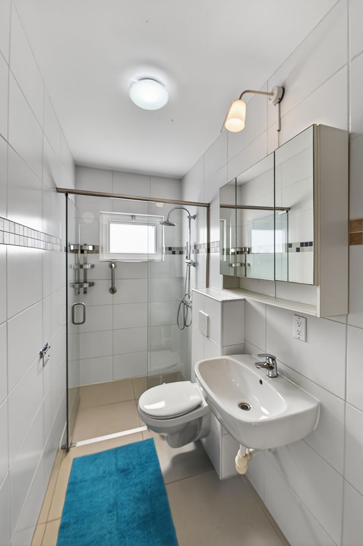 Modern white bathroom with walk-in shower, wall-mounted sink, toilet, mirrored cabinet, and teal bath mat.