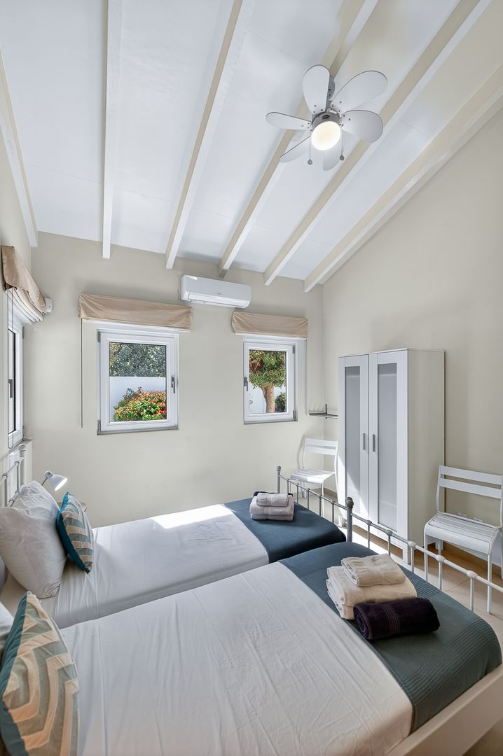 Airy twin bedroom with vaulted beamed ceiling, ceiling fan, two metal beds, white wardrobe, and garden windows.