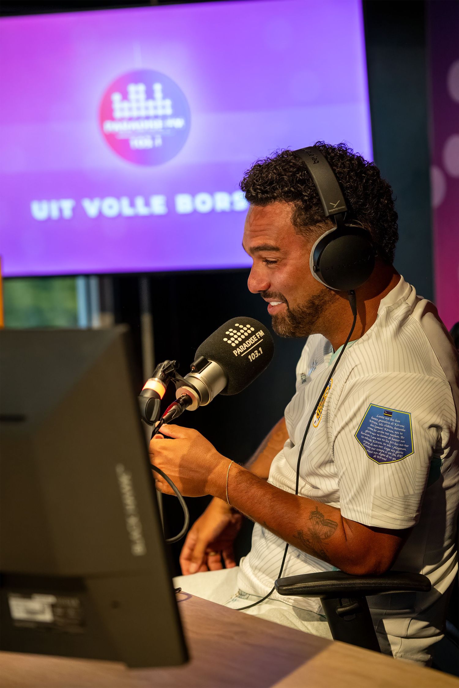 Radio host wearing headphones speaks into a microphone at a broadcast studio with a purple screen displaying Uit Volle Bor.