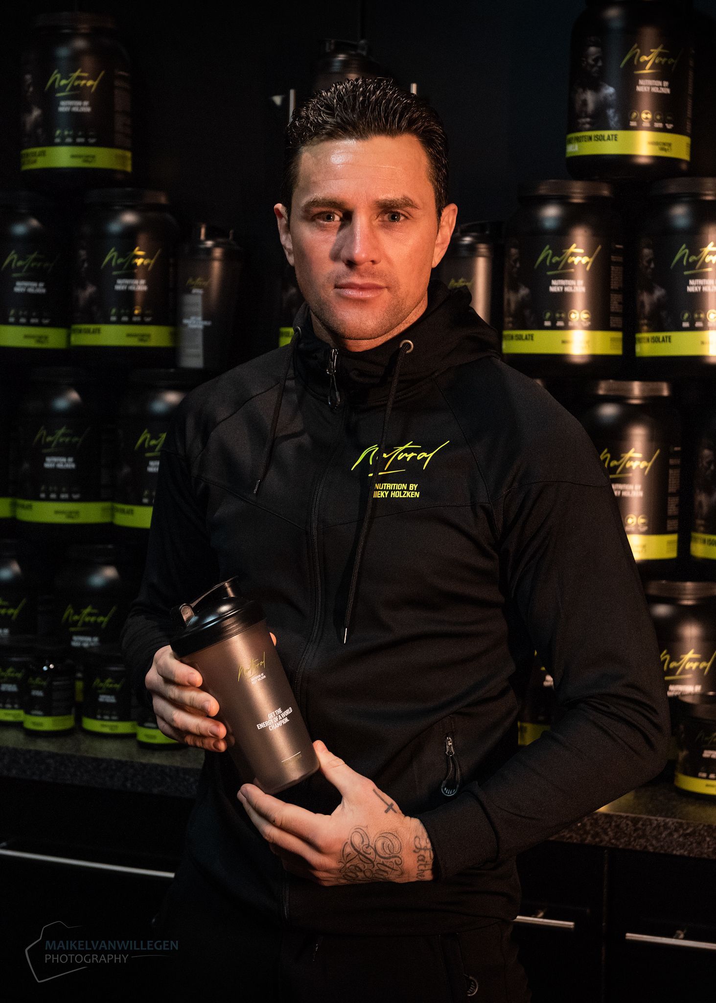 Person in black shirt holding and displaying a dark supplement container in a dimly lit retail environment.