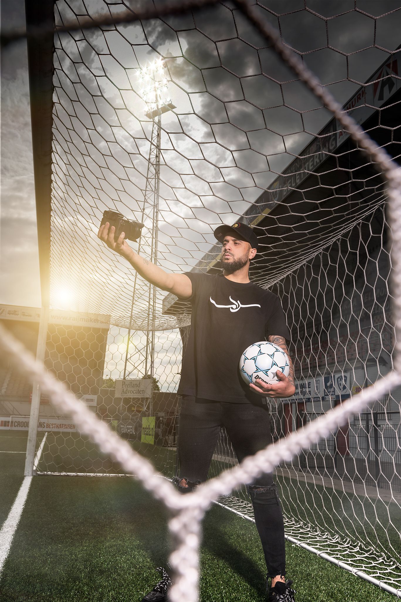 Soccer player in black athletic wear holds a ball while posing in front of a goal net during golden hour sunset.