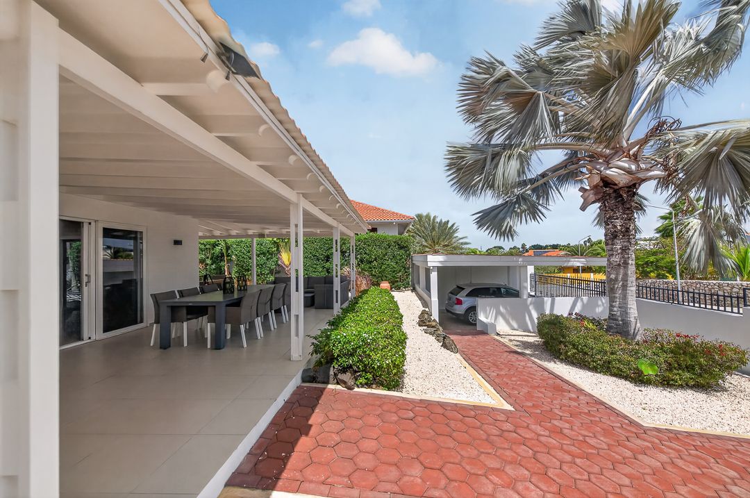 Spacious covered patio with outdoor dining area, lush hedges, palm tree, and brick driveway under a bright blue sky.