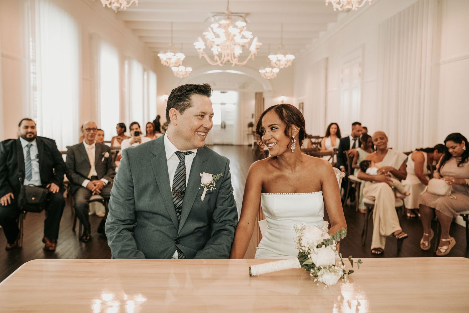 Elegant wedding reception with chandeliers and white decor features guests mingling in a bright ballroom.