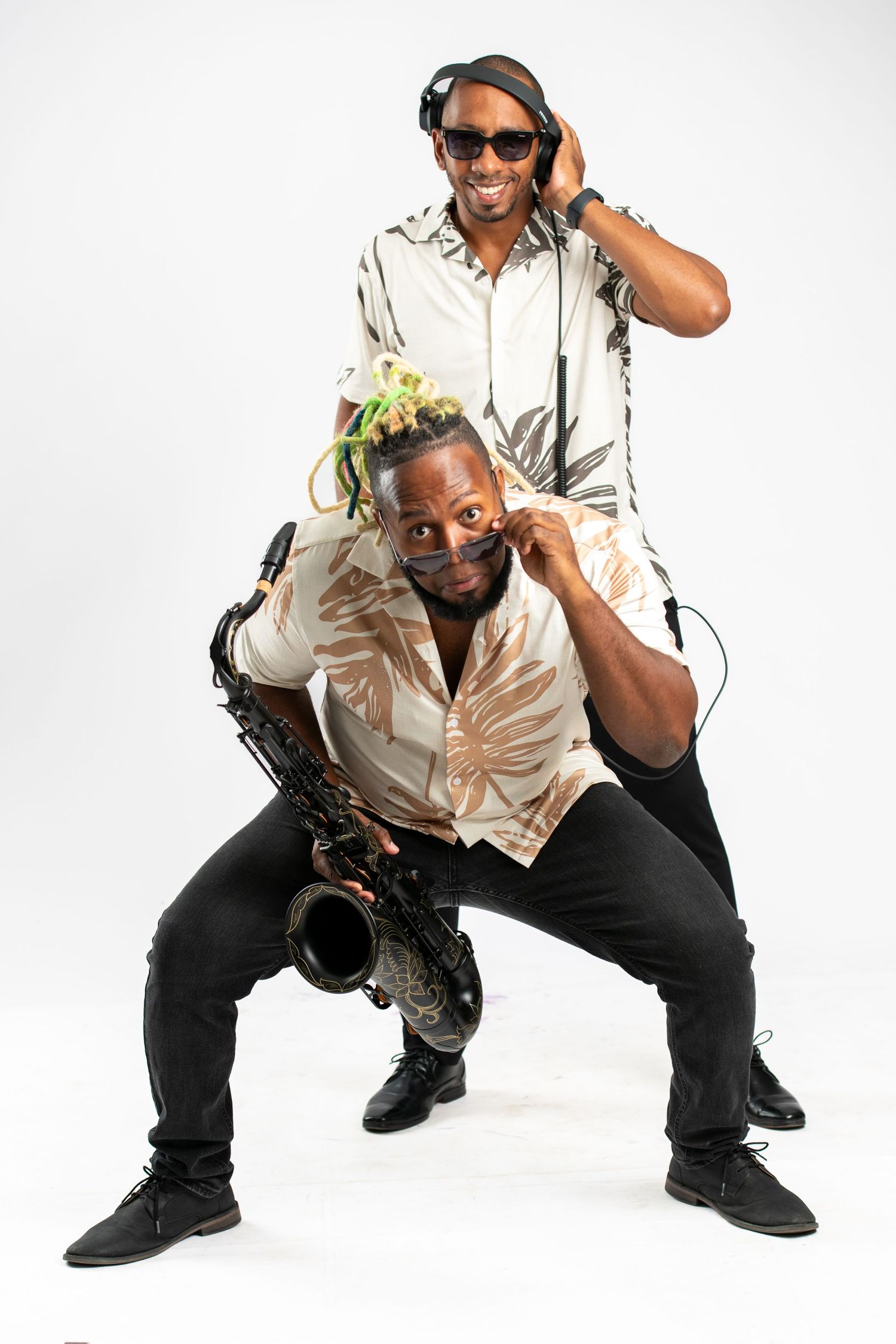 Two musicians pose playfully, one holding a saxophone and one wearing headphones, both in floral shirts on white background.