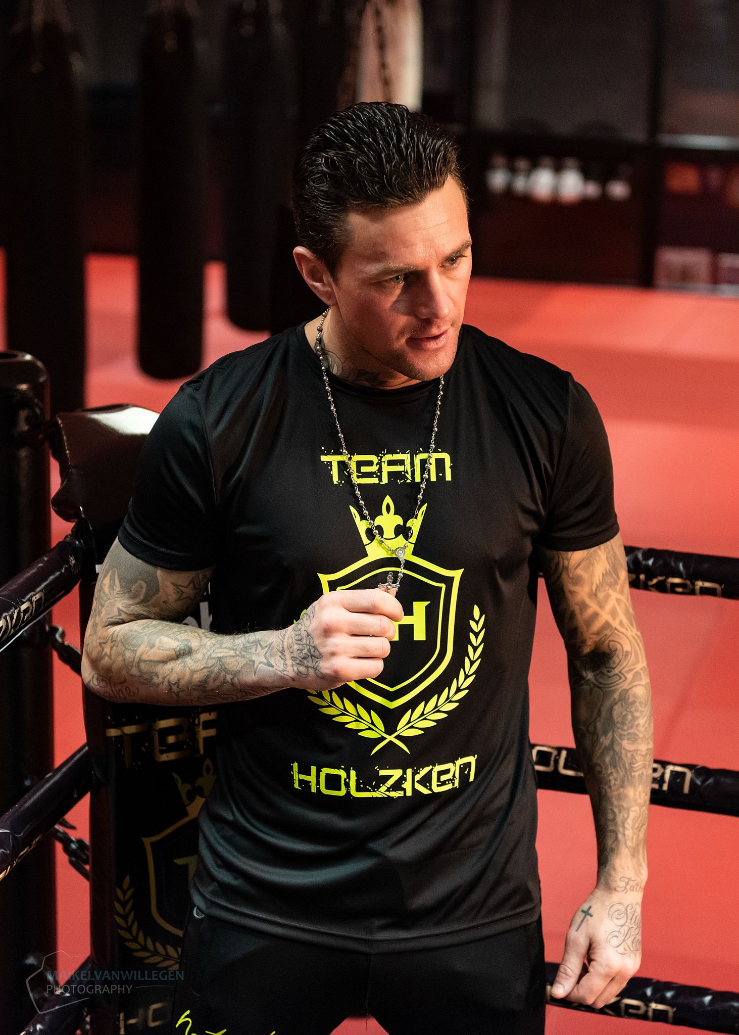 Athletic person in black Team Holzken t-shirt with tattoos stands near boxing ring ropes in red-lit gym setting.