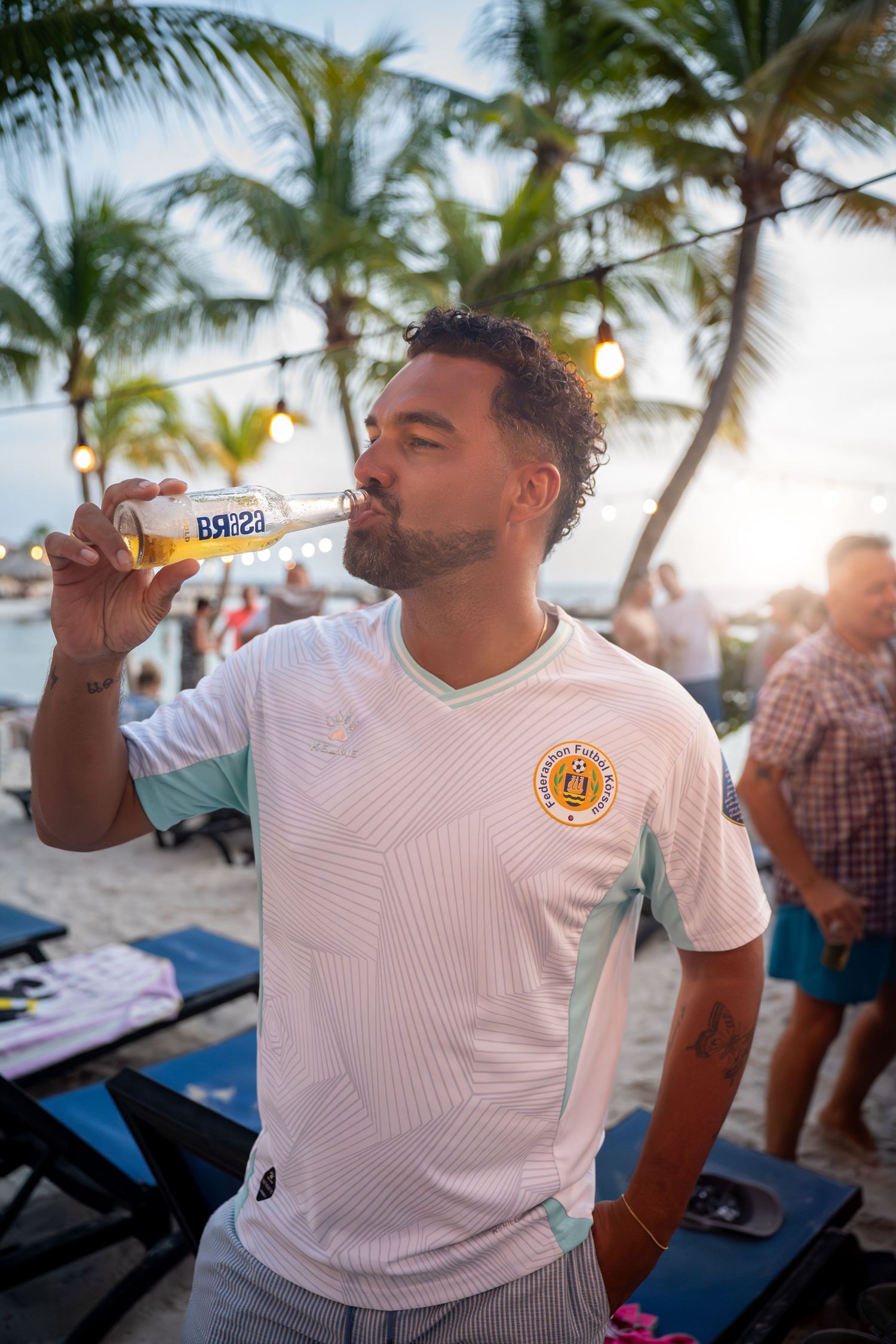 Man in white jersey drinking Brisa beer on a tropical beach at sunset with palm trees.