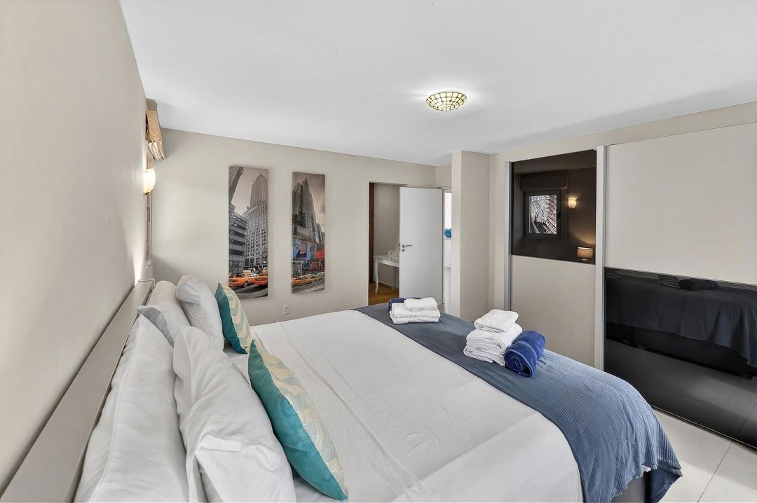 Modern hotel bedroom with king bed, teal accent pillows, city artwork, and ensuite bathroom in neutral tones.