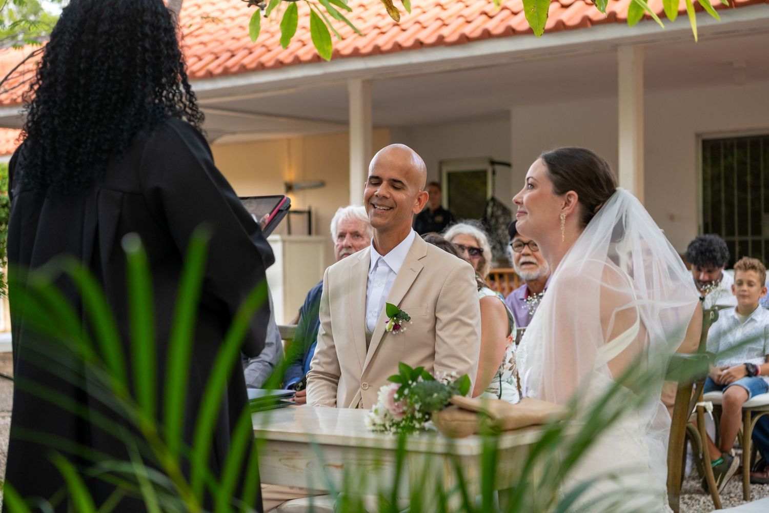 Wedding guests mingle and socialize during an outdoor reception under a covered patio with lush greenery.