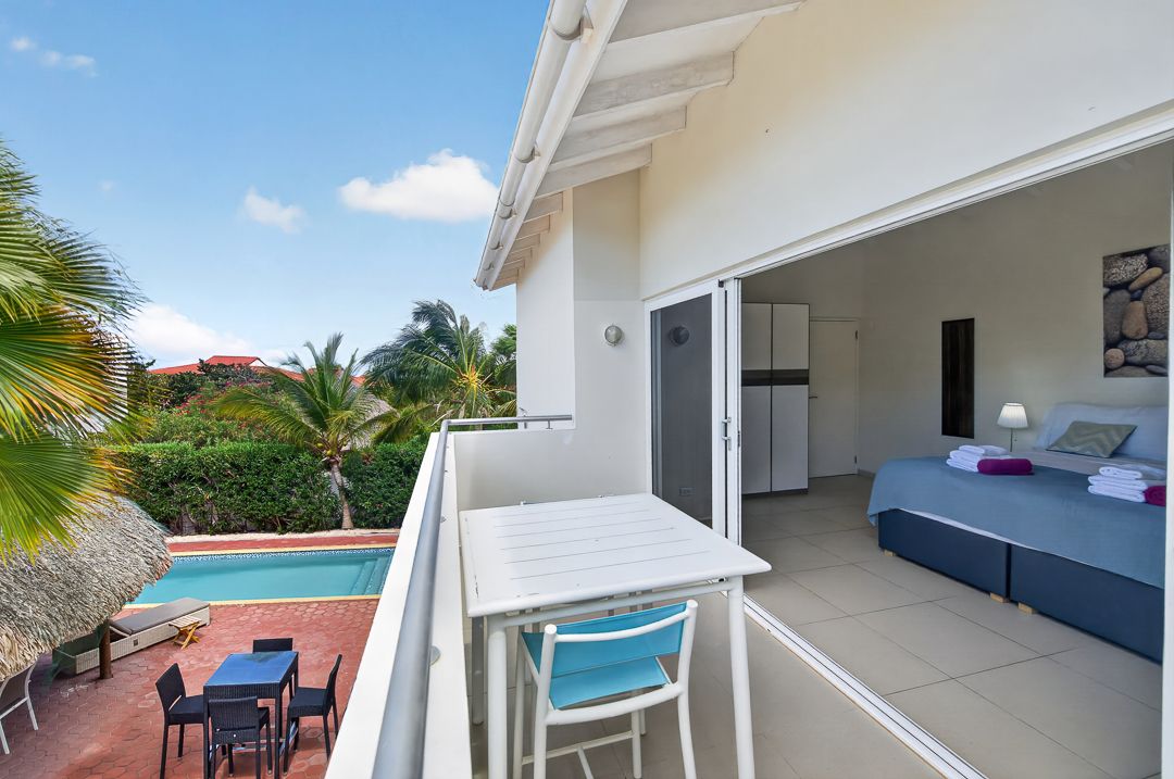 Bright balcony with white table, blue chairs, overlooking a pool and tropical garden, adjacent to a modern bedroom.