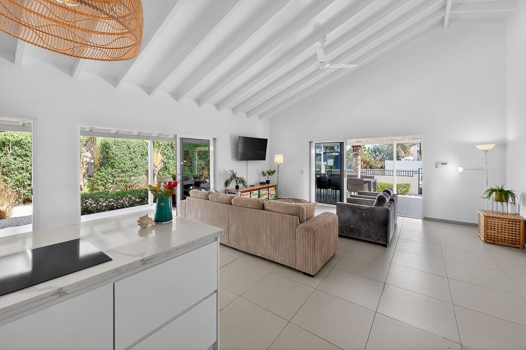 Bright open-plan living room with white beamed ceilings, tile floors, neutral sofas, and large windows overlooking lush greenery.