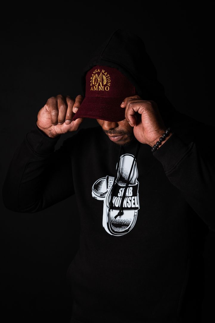 Portrait of person adjusting burgundy baseball cap while wearing black hoodie with sneaker graphic against dark backdrop.