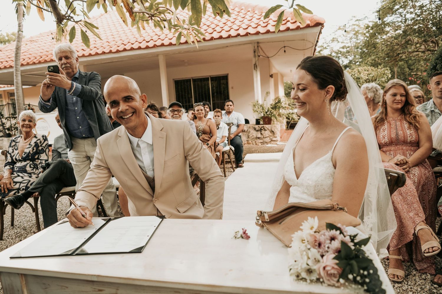 A joyful couple signs their marriage certificate at an outdoor ceremony with guests looking on from behind.