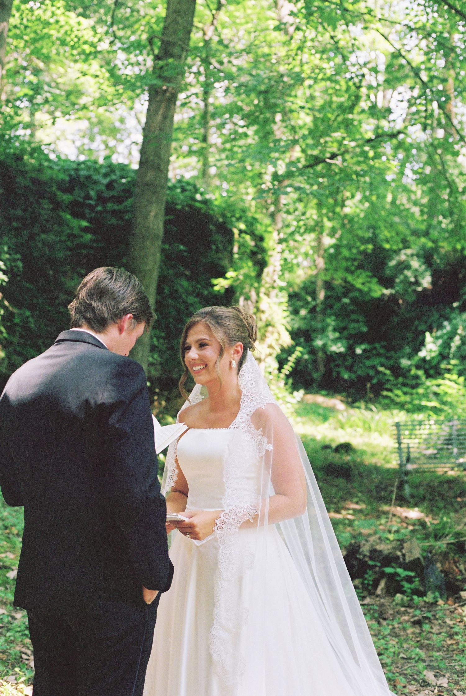 Smiling bride in white gown and veil exchanges vows with groom in suit surrounded by lush green trees.