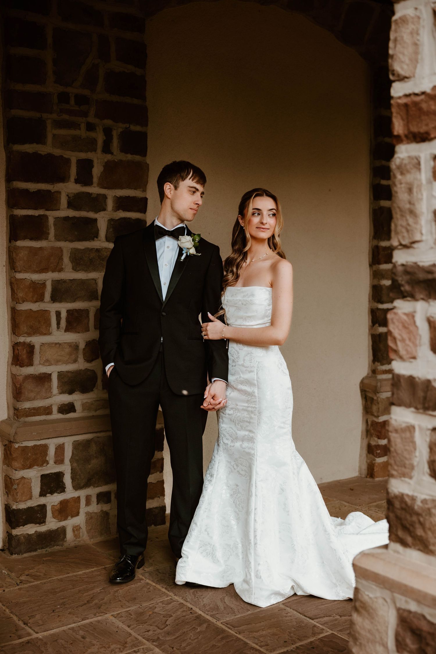 Bride in strapless mermaid gown and groom in black tuxedo pose together in a stone archway.