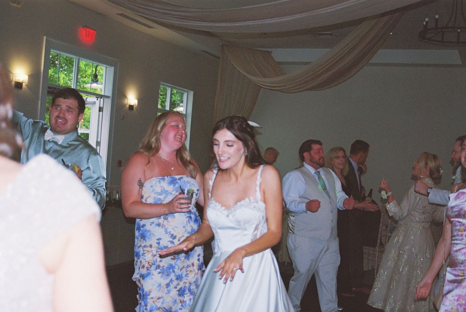Bride in white gown smiles while dancing with guests in elegant draped reception hall.