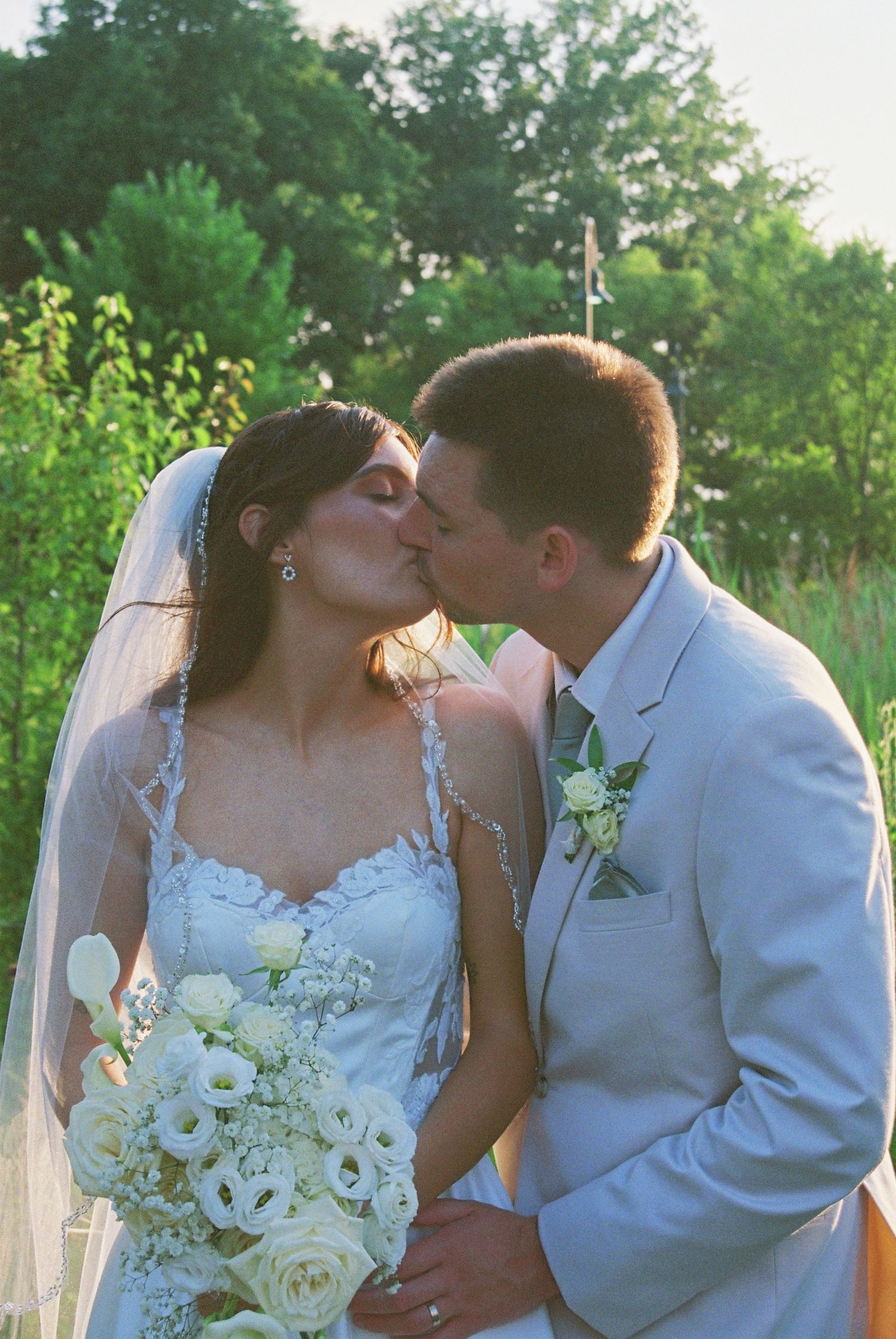 Bride and groom share a kiss outdoors, bride in lace gown holding white bouquet, groom in gray suit.