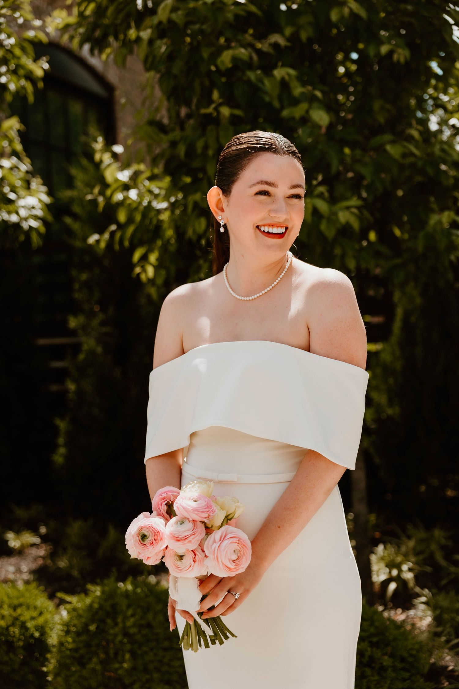 A bride in an off-shoulder white dress holds a pink rose bouquet while standing in a garden setting.