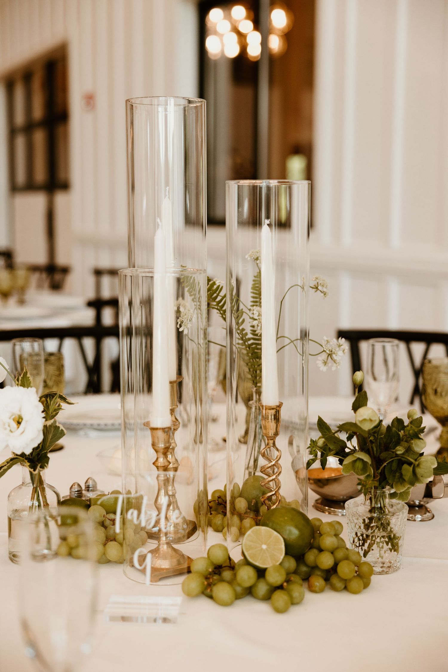 Elegant wedding table decor features tall glass candle holders, white flowers, and fresh grapes.