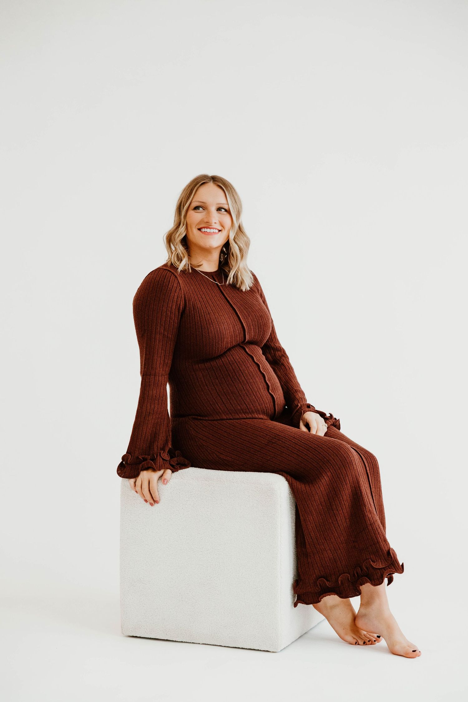 Person in brown knit dress poses on white cube in studio setting.