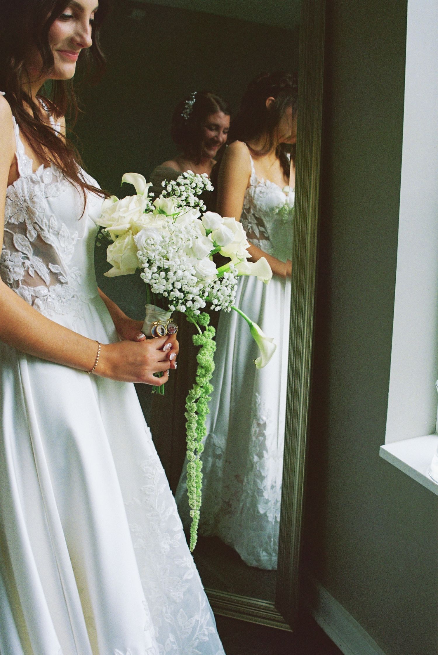 Bride in white lace gown holding cascading bouquet of white roses and calla lilies, reflected in a full-length mirror.