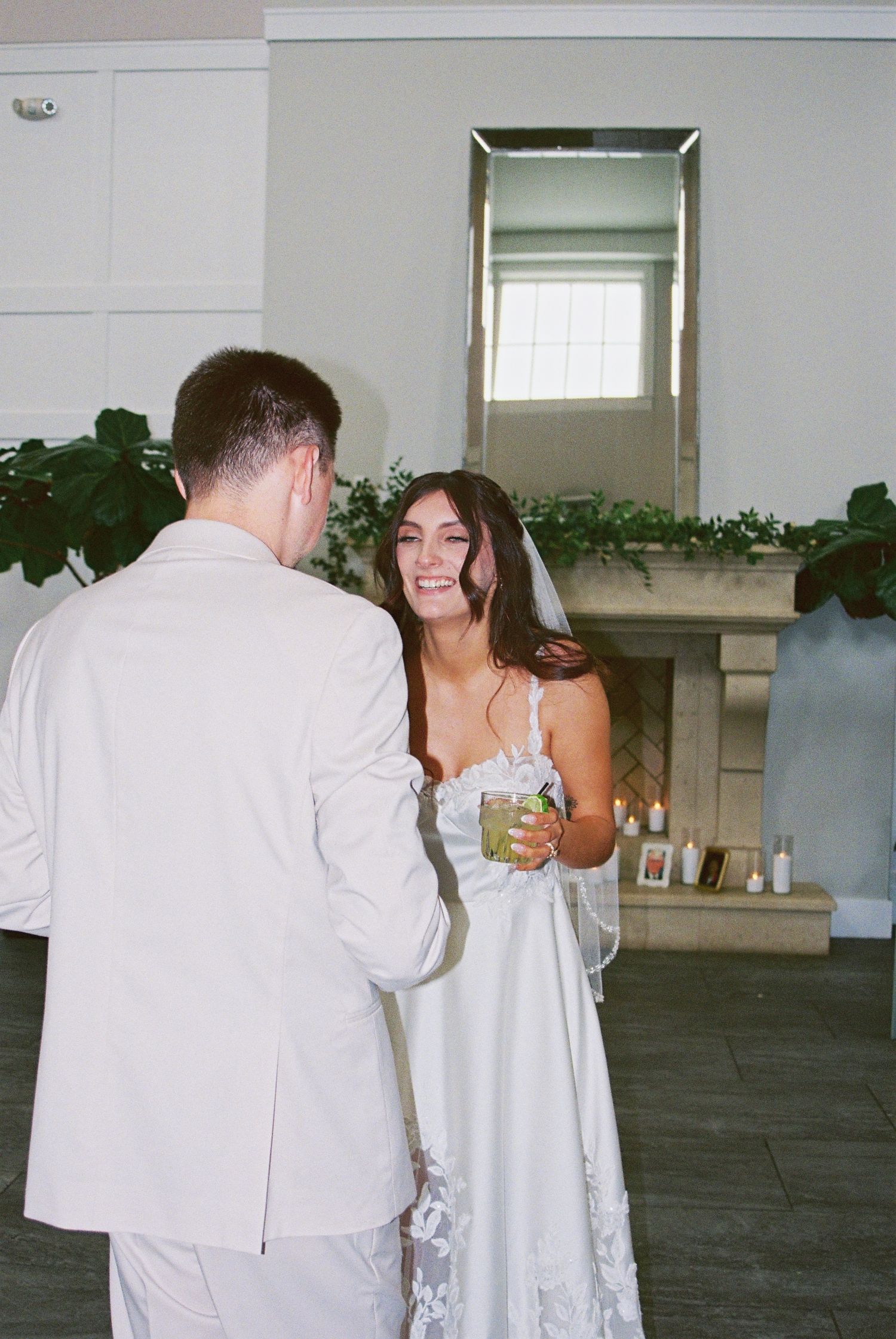 Smiling bride in lace gown holding a cocktail chatting with groom near a fireplace mantel.