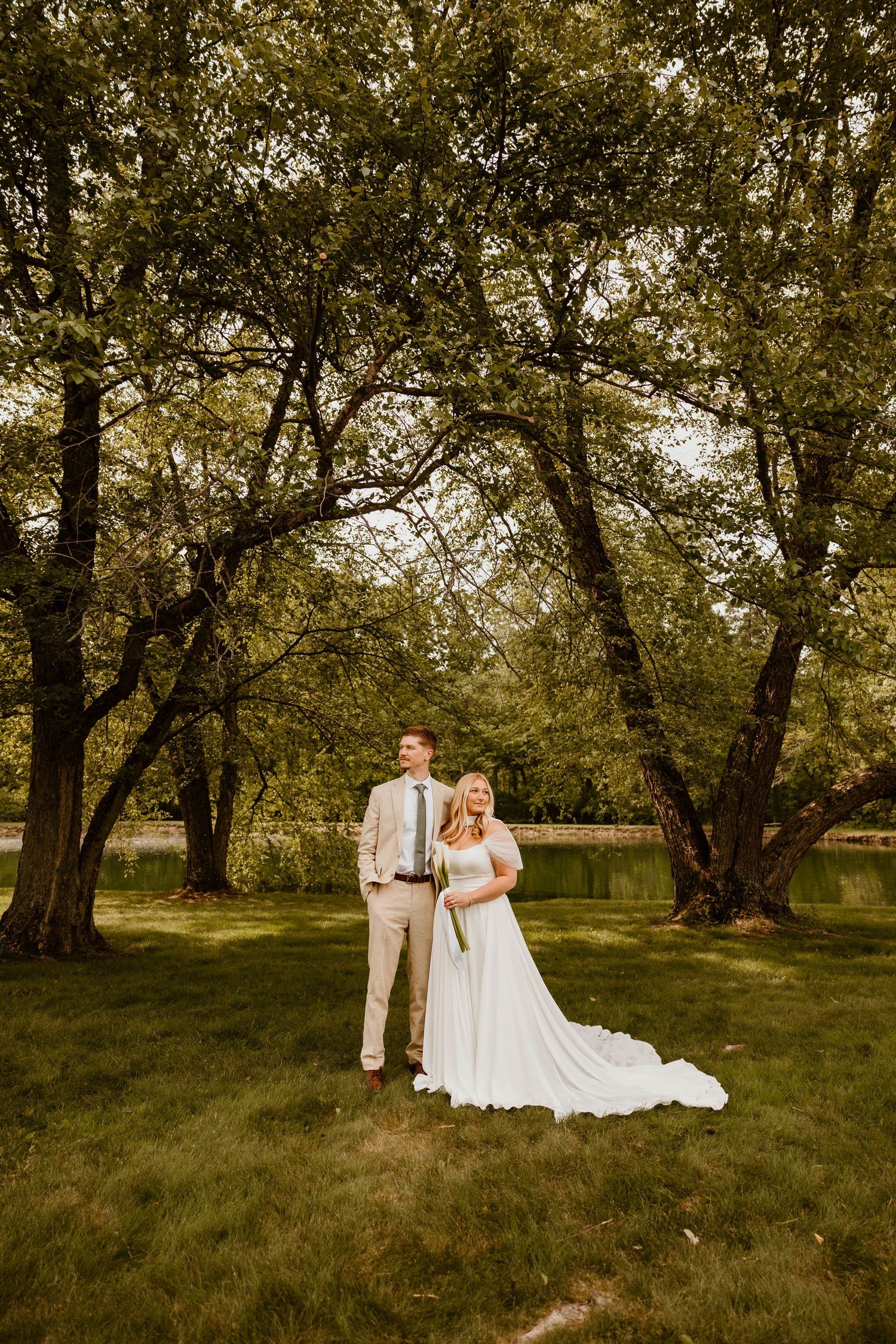 A wedding couple poses together on grass with tall trees in the background during golden hour.